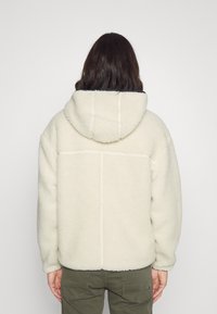 Chaqueta de forro polar color crema con capucha, que presenta una superficie suave y texturizada y un ribete en contraste. La parte posterior muestra detalles de costura y un corte relajado.