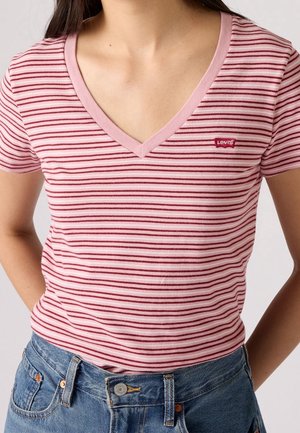 Osoba nosząca różowo-czerwony pasiasty T-shirt Levi's z dekoltem w serek, włożony do niebieskich dżinsów, ręce spoczywają z tyłu.