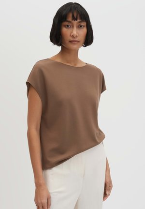 Frau mit kurzen schwarzen Haaren, die ein lockeres braunes ärmelloses Oberteil und weiße Hosen vor einem schlichten hellen Hintergrund trägt.