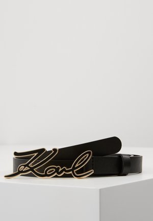 Czarny skórzany pasek z klamrą w kolorze złotym, w kształcie kursywnego napisu "Karl", umieszczony na białej powierzchni.