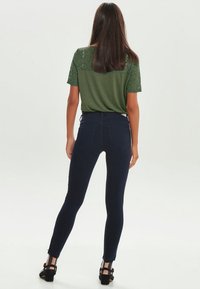 Camiseta de encaje y punto verde con mangas cortas, combinada con jeans ajustados de color azul oscuro. Los jeans tienen bolsillos traseros y una silueta ceñida.