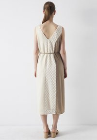 Vestido sin mangas beige con espalda en V, que presenta un panel de crema con patrón y un cinturón de tela en la cintura. Textura suave, diseño de largo hasta el suelo.