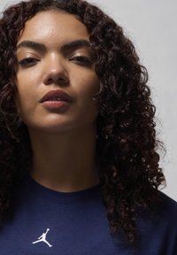 Mulher com cabelo encaracolado escuro, vestindo uma camisa azul marinho com um logotipo branco no peito, olhando confiantemente para a câmera contra um fundo simples.