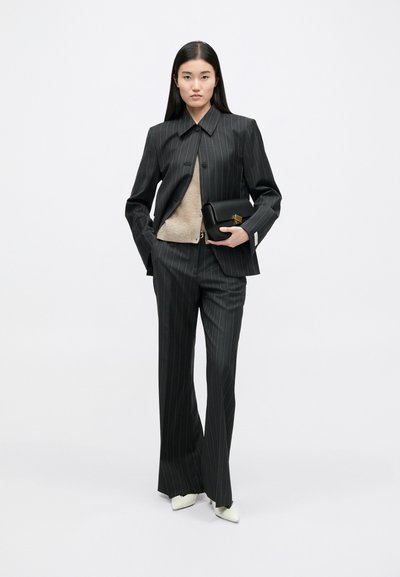 Zwart pinstripe pak met een getailleerd colbert en broek met wijde pijpen. Gecombineerd met een beige trui en een zwarte handtas. Witte puntige hakken.