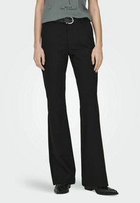 Pantaloni neri a palazzo con una texture liscia, dotati di passanti per cintura con una fibbia d'argento, abbinati a una camicia grigia. Scarpe nere con suola piatta.