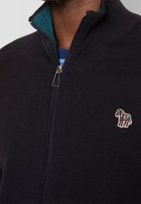 Hombre que lleva un suéter negro con cremallera, con un cuello interior teal y un pequeño parche de cebra a rayas de colores en el pecho.