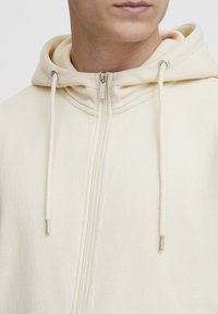 Felpa zip con cappuccio beige, morbido al tatto, con zip a contrasto e dettagli metallici sui lacci del cappuccio.
