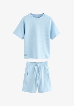 Dreng med kort hår iført lyseblå skjorte og shorts kryber sammen på gul gulv mod gul baggrund.