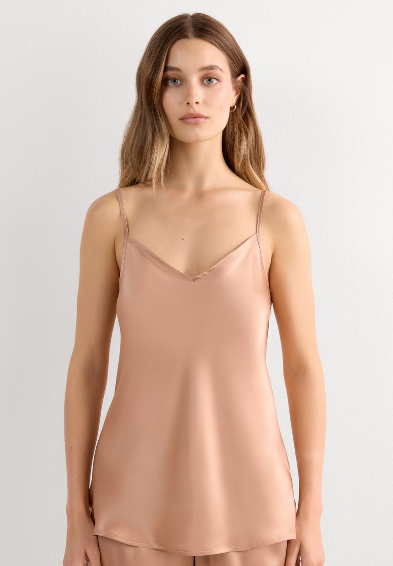 Young woman with long hair wears a beige satin camisole top with thin straps, standing against a plain light wall.
