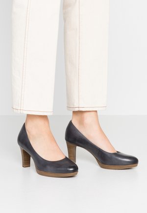 Persoon die witte broek en zwarte leren schoenen met houten zolen en middelhoog hak draagt, staand op een effen lichtgrijs oppervlak.