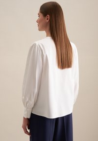 Blusa bianca con maniche lunghe a sbuffo, in tessuto morbido e scollatura rotonda. La parte posteriore ha una finitura liscia e dettagli sottili ai polsi.