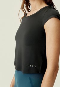 Černý zkrácený sportovní top s krátkými rukávy, vyrobený z hladké látky. Na dolním okraji je jemné logo. Nošeno s modrými legínami.