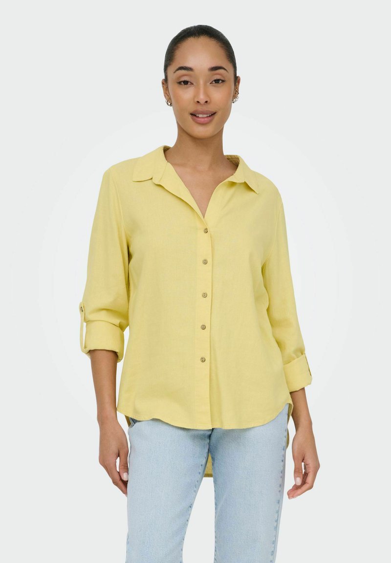 Femme portant une chemise jaune à boutons avec les manches retroussées et un jean bleu clair, debout devant un fond clair uni.