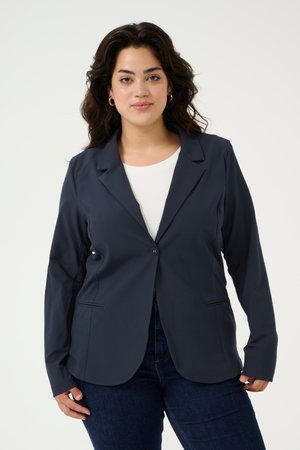 Blazer navy oscuro en tejido elástico, cierre de un solo botón, con solapas de muesca y bolsillos frontales. Lleva encima de una blusa blanca y combinado con jeans.