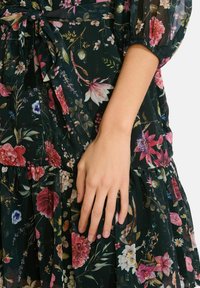 Dłoń spoczywająca na ciemnym materiale z kolorową sukienką w kwiaty, z różowymi, białymi i czerwonymi kwiatami oraz zielonymi liśćmi.