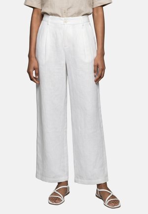 Donna che indossa pantaloni bianchi a vita alta, a gamba larga con pieghe, una camicia beige a maniche corte e sandali piatti bianchi con cinturini.