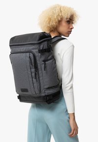 Zaino in tessuto a scacchi grigi e neri con molteplici tasche con zip, maniglia superiore e spallacci regolabili. Tessuto testurizzato con finitura opaca.