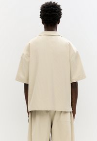 Beige rövid ujjú ing texturált anyagból, laza szabású, széles gallérral. Egyenes alja és minimális varrás részletei vannak.