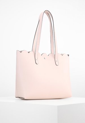 Lichtroze leren tote bag met een golvende bovenrand en dubbele schouderriemen op een wit voetstuk.