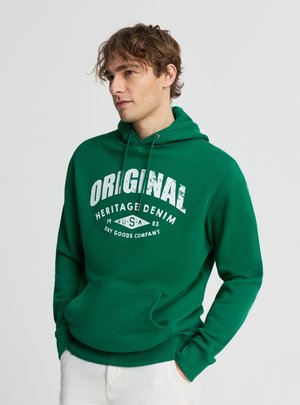 Giovane uomo con felpa verde con cappuccio con scritta bianca "ORIGINAL HERITAGE DENIM USA 1983", mani in tasca, guarda a sinistra.