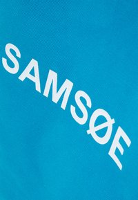 Τουρκουάζ ύφασμα με έντονα, λευκά γράμματα "SAM Sø E". Το ύφασμα έχει υφή, προσφέροντας μια ζωντανή και εντυπωσιακή εμφάνιση.