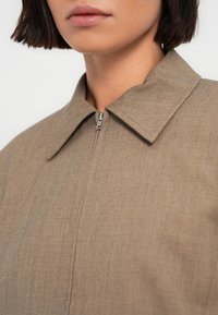 Κοντινό πλάνο ενός ατόμου που φοράει μπεζ παλτό με φερμουάρ και φαρδιά γιακά, εμφανίζοντας το κάτω μέρος του προσώπου και τον λαιμό.