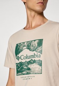 Beige bomulds T-shirt med et grønt bjerggrafik og teksten "Columbia Sportswear Company". Rund hals og korte ærmer.