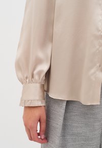 Blusa beige in satin con maniche lunghe gatherate e polsini arricciati, abbinata a tessuto grigio testurizzato. Superficie liscia con una leggera lucentezza.