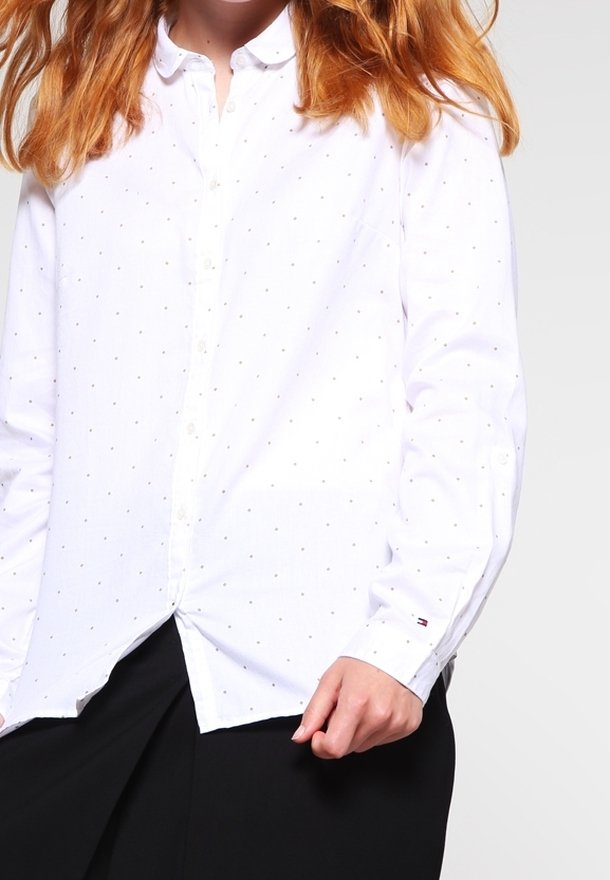 Camisa blanca de manga larga con pequeños puntos dorados dispersos. Cuenta con un cuello de botonadura y un bolsillo en el pecho. Textura de tela suave.
