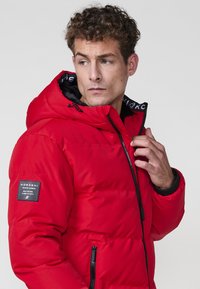 Chaqueta acolchada roja con capucha, que presenta un forro interior negro, cremallera frontal y parche con el logo en la manga izquierda. Diseño texturizado y cálido.
