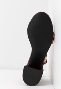 Semelle d’une sandale pour femme avec une semelle inférieure noire texturée et des brides marron à motif écaille de tortue sur fond blanc.