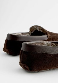 Des pantoufles en daim marron avec des accents en cuir et une doublure texturée, présentant un profil bas et un design arrondi. Des semelles épaisses assurent la stabilité.