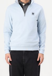 Uomo che indossa una felpa leggera azzurra con zip a metà con logo sul petto, sopra una polo grigia scura e jeans neri, in piedi con le braccia rilassate.