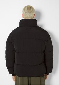 Svart puffjacka med hög krage och quiltat mönster. Har en slät yta och elastiska ärmslut. Baksidan visar sömmarnas detaljer.