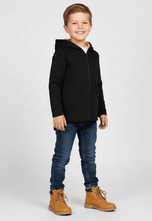 Uśmiechnięty chłopiec stojący, ubrany w czarną bluzę na zamek, niebieskie jeansy i beżowe buty sznurowane na jasnym, jednolitym tle.