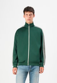 Veste zippée verte avec un col haut, ornée de bandes blanches contrastantes le long des épaules. Fabriquée en tissu texturé, coupe décontractée.