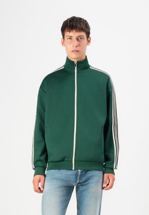 Grüne Zip-Jacke mit hohem Kragen, mit kontrastierenden weißen Streifen an den Schultern. Hergestellt aus strukturiertem Stoff, mit lockerer Passform.