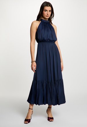 Vrouw draagt een mouwloze marineblauwe halter-neck maxi-jurk met plooien, enkellaarsjes met riempje en een gouden armband, staand tegen een effen achtergrond.