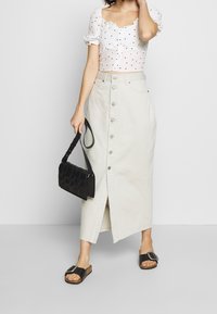 Kvinna iklädd en vit topp med prickar, en beige knäppt midiklänning, svarta slip-in sandaler och bärande på en svart vävd handväska.