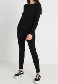 Camicia nera a maniche lunghe, abbinata a jeans skinny neri e sneaker Vans nere, in piedi su uno sfondo bianco uniforme.