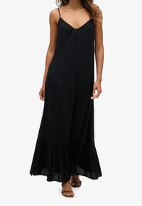 Svart maxiklänning med tunna stroppar, V-ringning och strukturerad tyg. Har en utsvängd nederkant med ett volangdetalj längst ner.