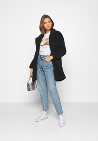 Manteau long noir et texturé sur un t-shirt graphique, associé à un jean taille haute bleu clair et des baskets blanches. Tenant un petit sac à motifs.