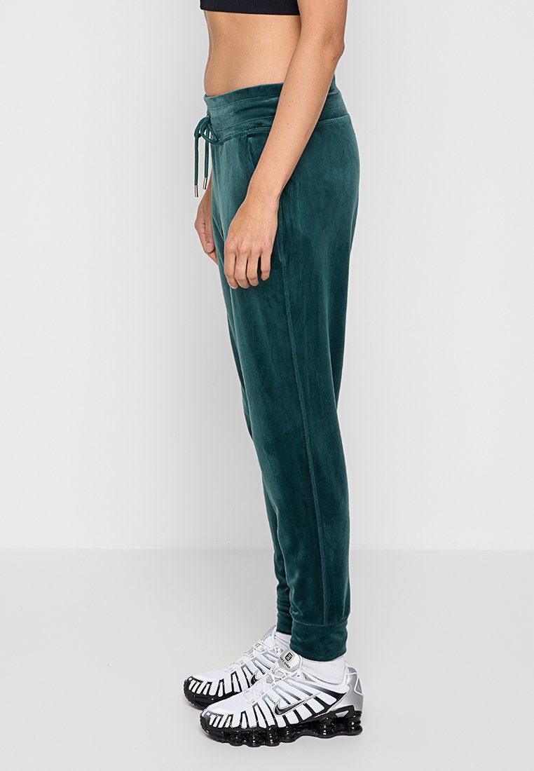 Kobieta ubrana w ciemnozielone dresowe spodnie z weluru oraz białe i czarne sneakersy, stojąca bokiem na tle gładkiej białej ściany.