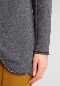 Top de manga larga gris con textura, con un dobladillo redondeado y bordes deshilachados, usado sobre pantalones de color mostaza, mostrando un diseño relajado.