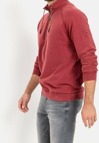 Polaire rouge à col demi-zip, avec cordons de serrage noirs et jaunes et poignets côtelés. Portée avec un jean gris.