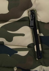 Tessuto in puffa camouflage con tonalità di verde e beige, dotato di una cerniera nera con marchio e dettagli di cucitura a contrasto.