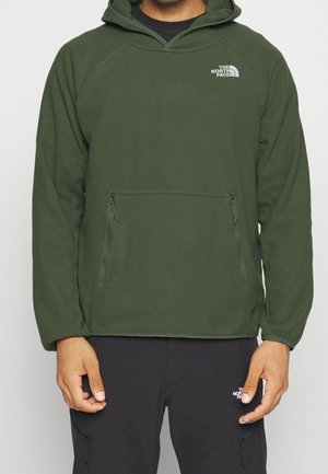 Mies, jolla on päällään oliivinvihreä fleece-huppari ja mustat housut, molemmissa The North Face -logo, seisoo yksinkertaisen taustan edessä.