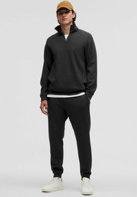 Felpa nera con zip e collo alto, abbinata a pantaloni jogger neri e sneakers color crema. Texture liscia, vestibilità comoda, design minimalista.