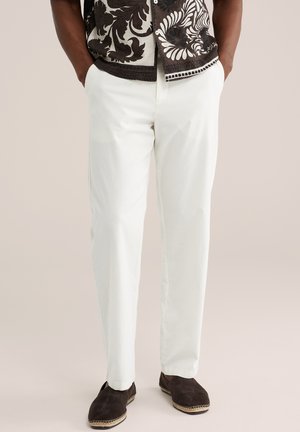 Fehér nadrágot, sötétbarna kényelmes cipőt és fekete-fehér mintás rövid ujjú inget viselő férfi, kezét zsebében tartva.