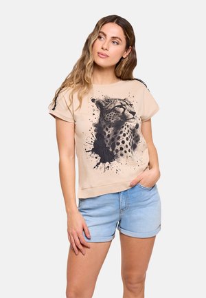 Mujer con el pelo largo que lleva una camiseta beige con un gráfico de guepardo y pantalones cortos de mezclilla azul claro enrollados, de pie con una mano en el bolsillo.